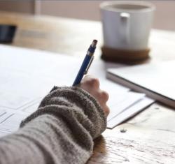 Photo d'une main écrivant sur un cahier. Une tasse sur la table au second plan.